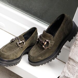 May include: A pair of olive green suede loafers featuring a thick black lug sole and a metallic horsebit detail across the vamp. The shoes have a rounded toe, visible stitching along the upper, and a dark interior lining for a structured appearance.