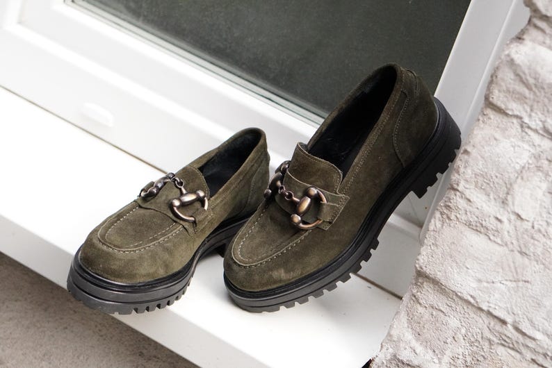 May include: A pair of olive green suede loafers featuring a thick black lug sole and a metallic horsebit detail across the vamp. The shoes have a rounded toe, visible stitching along the upper, and a dark interior lining for a structured appearance.