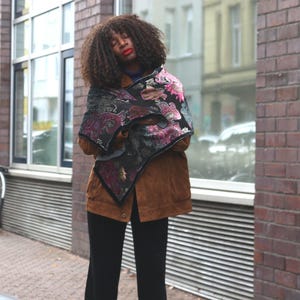 Op de afbeelding: Een persoon draagt een bruine suède jas, een zwarte broek en een bloemen sjaal met roze en paarse accenten. De persoon staat voor een bakstenen gebouw met een geparkeerde fiets.