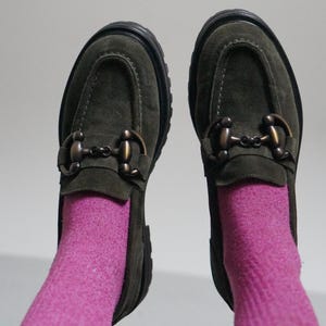 May include: A pair of olive green suede loafers featuring a dark bronze-toned metal horsebit detail across the vamp and a thick black lug sole. The shoes are worn with bright pink socks that extend upward from the opening of each loafer.