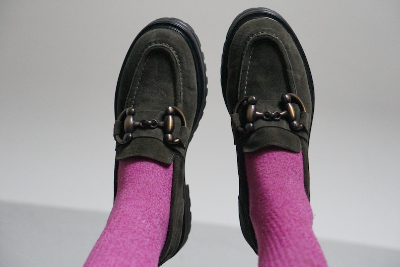 May include: A pair of olive green suede loafers featuring a dark bronze-toned metal horsebit detail across the vamp and a thick black lug sole. The shoes are worn with bright pink socks that extend upward from the opening of each loafer.