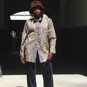 Könnte beinhalten: Eine knöpfbare Jacke mit Schlangenmuster in Beige-, Braun- und Blautönen. Die Jacke wird mit einer dunkelblauen Samthose und einem braunen Fischerhut kombiniert. Das Outfit wird in einer gut beleuchteten Umgebung im Freien getragen.