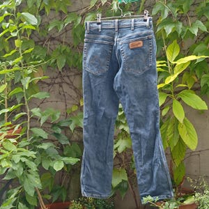 Pode incluir: Jeans azuis de ganga pendurados num cabide. Os jeans t&ecirc;m um design cl&aacute;ssico de cinco bolsos, com um remendo de couro castanho no bolso de tr&aacute;s. Os jeans est&atilde;o desbotados e desgastados, com um ligeiro alargamento na parte inferior.