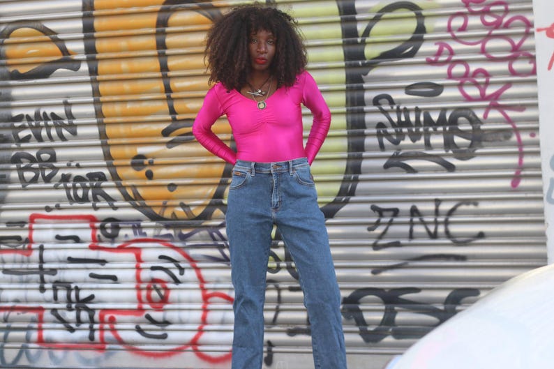 Pode incluir: Uma pessoa vestindo um top rosa brilhante de manga comprida e jeans azuis. O fundo &eacute; uma superf&iacute;cie met&aacute;lica coberta de grafite com tinta amarela, laranja e preta. A pessoa est&aacute; usando um colar.