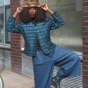 May include: A person wearing a green, blue, and white plaid jacket with a black turtleneck, blue denim culottes, and a plaid hat. The person is wearing black boots. A bicycle is in the background.
