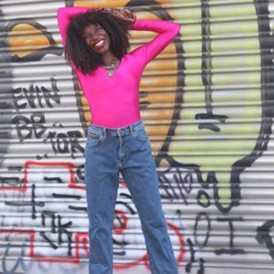 Pode incluir: Uma pessoa vestindo uma blusa rosa brilhante de manga comprida e jeans azuis. O fundo &eacute; uma persiana de metal coberta de grafite. A pessoa est&aacute; sorrindo e com os bra&ccedil;os levantados acima da cabe&ccedil;a.