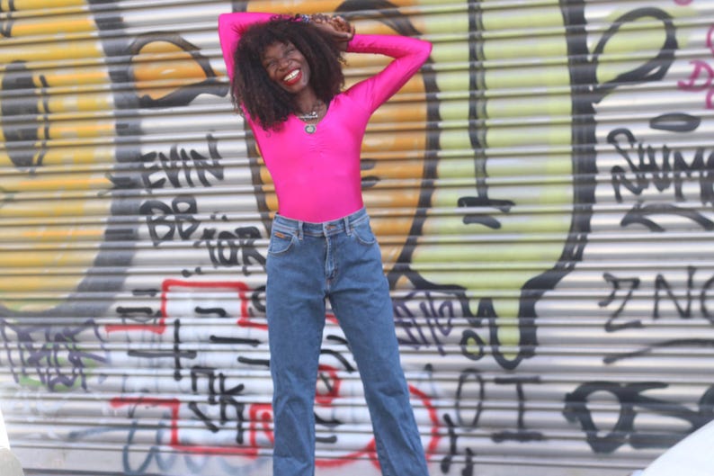 Pode incluir: Uma pessoa vestindo uma blusa rosa brilhante de manga comprida e jeans azuis. O fundo &eacute; uma persiana de metal coberta de grafite. A pessoa est&aacute; sorrindo e com os bra&ccedil;os levantados acima da cabe&ccedil;a.