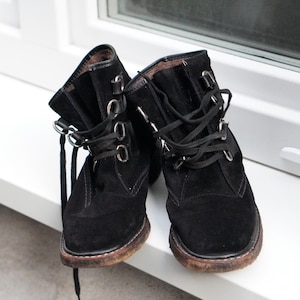 May include: A pair of black suede ankle boots with laces and metal eyelets. The boots have a worn, brown sole and are displayed on a white surface near a window.