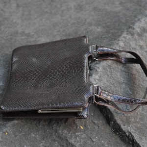 Può includere: Una borsa a mano marrone scuro con un motivo testurizzato in pelle di serpente. La borsa presenta una forma rettangolare, una chiusura con telaio in metallo sul bordo superiore e due manici abbinati fissati con hardware in metallo sul lato.