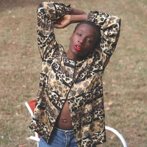 Op de afbeelding: Een jasje met knopen en een luipaardprint in bruin, zwart en beige tinten. De jas is open en onthult een glimp van de buik van de persoon. De persoon draagt een blauwe spijkerbroek en heeft de armen boven het hoofd geheven.