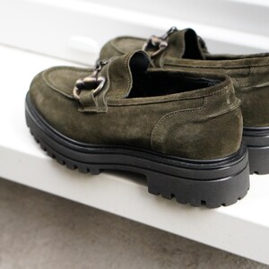 May include: A pair of olive green suede loafers featuring a dark metal horsebit detail across the vamp. The shoes have a thick, black, lugged rubber sole with a deep tread pattern, providing a chunky silhouette and a rounded toe design.