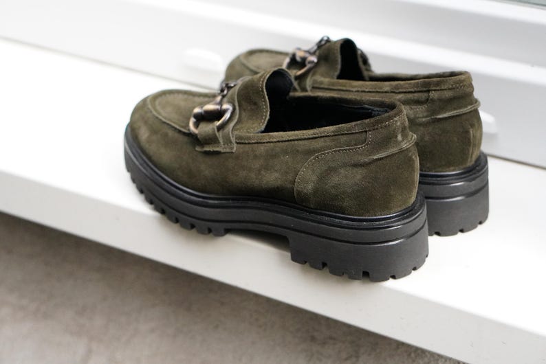 May include: A pair of olive green suede loafers featuring a dark metal horsebit detail across the vamp. The shoes have a thick, black, lugged rubber sole with a deep tread pattern, providing a chunky silhouette and a rounded toe design.