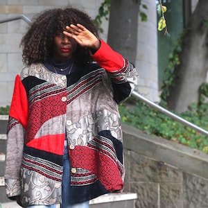 May include: A long-sleeved jacket featuring a patchwork design with panels of red, black, and grey fabric. The garment includes intricate embroidered patterns, a round neckline, and a front button closure with metallic buttons.