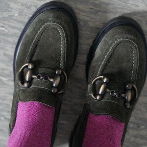 May include: A pair of olive green suede loafers featuring a prominent bronze-toned metal horsebit detail across the top. The shoes have a thick black sole and are worn with bright magenta socks, showcasing a textured finish and visible stitching.