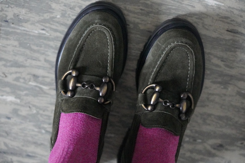 May include: A pair of olive green suede loafers featuring a prominent bronze-toned metal horsebit detail across the top. The shoes have a thick black sole and are worn with bright magenta socks, showcasing a textured finish and visible stitching.