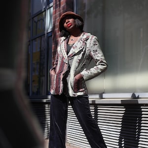 May include: A patterned blazer with a floral design in shades of green, brown, and cream. The blazer has a striped panel on the left side and is paired with dark velvet pants. A brown hat completes the look.
