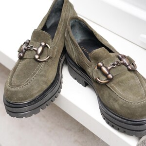 May include: A pair of olive green suede loafers featuring a thick black lug sole and a metallic bronze-toned horsebit detail across the top of the vamp. The shoes have a rounded toe and visible stitching along the front edge of the upper.