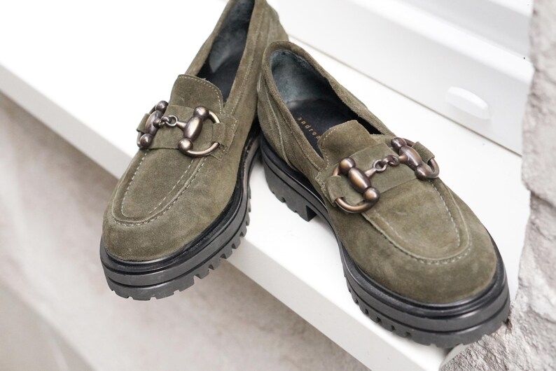 May include: A pair of olive green suede loafers featuring a thick black lug sole and a metallic bronze-toned horsebit detail across the top of the vamp. The shoes have a rounded toe and visible stitching along the front edge of the upper.