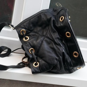 Può includere: Borsa a tracolla in pelle nera con occhielli e finiture dorate. La borsa presenta un design trapuntato e una lunga tracolla regolabile. La borsa è su una superficie bianca.