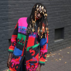 Könnte beinhalten: Ein langer, farbenfroher Cardigan mit einem auffälligen geometrischen Design. Der Pullover zeigt eine Mischung aus leuchtenden Farben, darunter Pink, Rot, Blau, Grün und Schwarz. Zahlen und Text sind in das Design integriert. Der Cardigan wird über einem dunklen Outfit getragen.