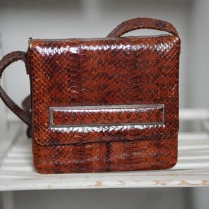 May include: A vintage brown snakeskin handbag with a rectangular shape and a silver-toned metal accent. The bag has a top handle and a shoulder strap. The bag is sitting on a white wooden shelf.