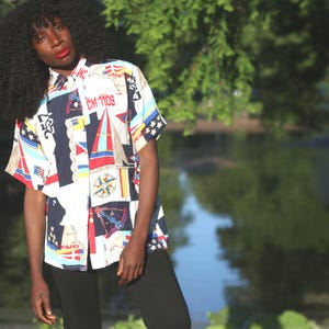 May include: Short-sleeved button-up shirt with a nautical-themed print featuring sailboats, flags, and geometric shapes in red, white, and navy blue. The shirt has a relaxed fit and is worn with black pants.