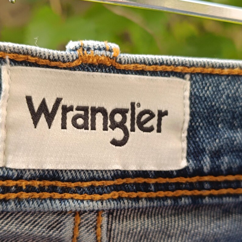Pode incluir: Close-up de uma pe&ccedil;a de roupa de ganga com a marca "Wrangler" impressa em preto numa etiqueta branca. A etiqueta &eacute; cosida ao tecido de ganga azul com linha laranja.
