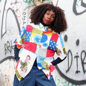 Peut inclure: Une chemise patchwork colorée à manches courtes, avec des panneaux rouges, bleus, jaunes et blancs. Portée sur un col roulé noir et associée à un pantalon en jean bleu. Le fond présente des graffitis.