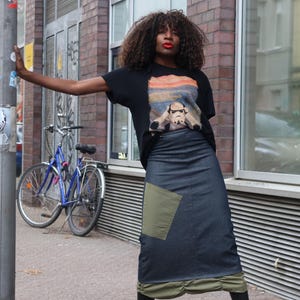 Op de afbeelding: Zwart T-shirt met een Stormtrooper-afbeelding, gecombineerd met een lange spijkerbroekrok met een olijfgroene patch. Op de achtergrond is een blauwe fiets te zien.