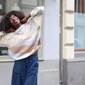 Può includere: Una persona indossa un maglione color crema con un motivo colorato a righe marroni, blu e beige. La persona indossa anche pantaloni di jeans blu. Lo sfondo comprende un edificio con una finestra e una porta.