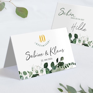 May include: Two white folded cards with text and botanical accents. One card reads "Sabine & Klaus" with the number "40" in gold. The other card says "Schön, dass du da bist Hilde". Both cards have green eucalyptus leaf designs.