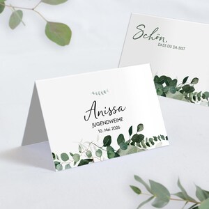 Op de afbeelding: Twee witte gevouwen kaarten met groene eucalyptusbladaccenten. Eén kaart leest "Anissa, JUGENDWEIHE, 10. Mai 2026" in zwart schrift. De andere kaart leest "Schön, dass du da bist" in groen schrift. Eucalyptus takken op de achtergrond.