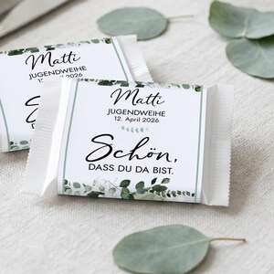 May include: Two wrapped candy bars with the text "Matti Jugendweihe 12. April 2026 Schön, dass du da bist." in black script. The wrappers have a white background with green floral accents. A knife and plate are in the background.