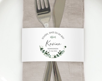 May include: A white plate with a folded beige linen napkin, a fork, and a knife. A white paper band with text and a green floral design encircles the napkin. The text includes the name "Karina" and the date "10. Mai 2020".