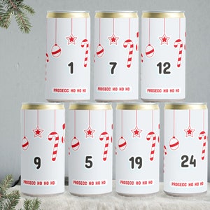Op de afbeelding: Zeven witte aluminium blikjes met gouden deksels, elk met een ander nummer van 1 tot 24, een rode ster, een rode snoepstok en een rode kerstbal. De blikjes hebben de tekst "PROSECC HO HO HO" erop gedrukt.
