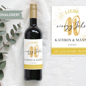 Op de afbeelding: Een donkere wijnfles met een gouden dop en een etiket met de tekst "LIEBE vierzig Jahre KATHRIN & MÄNNE 17.09.2025 EIN GOLDENER TROPFEN." Een bijpassende kaart staat naast de fles. Eucalyptus takken staan op de achtergrond.