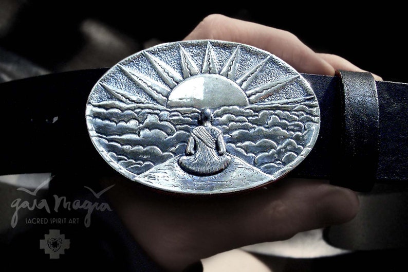May include: A silver belt buckle with a detailed design of a person sitting in meditation with a sunrise behind them. The buckle is oval shaped and has a textured surface.
