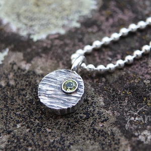 May include: A silver necklace with a round pendant featuring a textured surface and a small green gemstone. The necklace is made of silver ball chain.