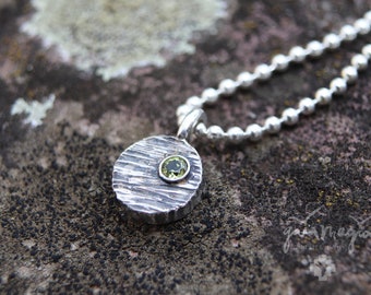 Op de afbeelding: Een zilveren ketting met een ronde hanger met een getextureerd oppervlak en een kleine groene edelsteen. De ketting is gemaakt van een zilveren bolketting.