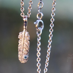 May include: A gold-colored feather pendant necklace with a small blue gemstone at the base of the feather. The necklace chain is a delicate link style. The background is a soft, blurred green.