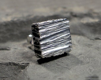 May include: A square silver earring with a textured, wood-grain-like surface. The earring is set against a dark gray, textured background. The design is minimalist and modern, suitable for everyday wear.