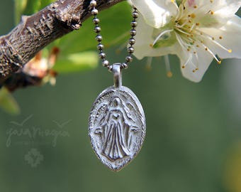 Op de afbeelding: Zilveren ovale hangerketting met een religieuze figuur. De hanger hangt aan een kogelketting. De achtergrond is een wazige afbeelding van een tak met witte bloemen en groene bladeren.