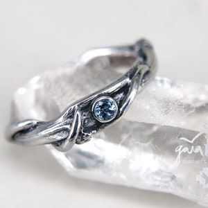 Könnte beinhalten: Ein silberfarbener Ring mit einem von der Natur inspirierten Design. Der Ring hat ein strukturiertes, astähnliches Band und einen kleinen, runden, hellblauen Edelstein. Der Ring wird auf einer transparenten, kristallinen Oberfläche präsentiert.