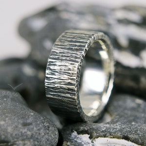 Könnte beinhalten: Ein silberner Ring mit einer strukturierten, rindenartigen Oberfläche. Der Ring ist vor einem verschwommenen Hintergrund aus grauen Steinen platziert. Das Design des Rings weist eine raue, organische Textur auf, die ihm ein einzigartiges, handgefertigtes Aussehen verleiht.