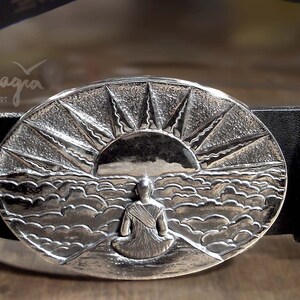May include: A silver belt buckle with a detailed design of a person sitting in front of a sunrise over a sea of clouds. The buckle is oval shaped and has a textured surface. The text "gaia magia" is engraved on the buckle.