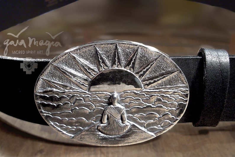 May include: A silver belt buckle with a detailed design of a person sitting in front of a sunrise over a sea of clouds. The buckle is oval shaped and has a textured surface. The text "gaia magia" is engraved on the buckle.