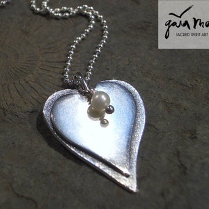 Op de afbeelding: Een zilveren ketting met een dubbele hart hanger en een parel accent. De hanger is gelaagd en heeft een textuur. De ketting is op een zilveren ketting.