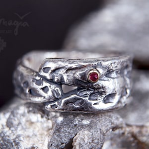 Puede incluir: Un anillo de plata con un diseño de serpiente texturizado y un ojo de piedra roja. El anillo está colocado sobre un fondo de piedra gris.