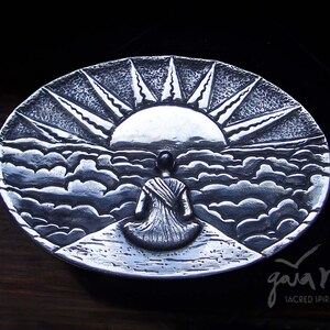 May include: A silver metal dish with a raised design of a person sitting in front of a sunrise over clouds. The dish is signed "Gaia Magia Sacred Spirit Art".