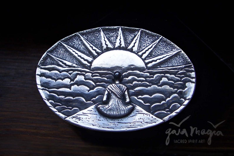 May include: A silver metal dish with a raised design of a person sitting in front of a sunrise over clouds. The dish is signed "Gaia Magia Sacred Spirit Art".